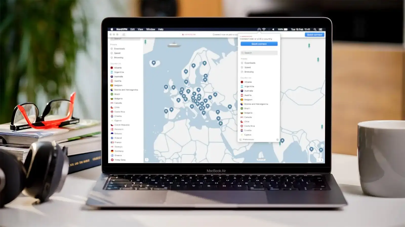 A MacBook Air displaying the NordVPN interface with a map of Europe and available server locations. The laptop is on a desk next to books, a pair of glasses, and a cup of coffee, suggesting a cozy and secure workspace.